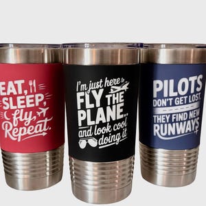 May include: Three stainless steel travel mugs with black lids. One mug has a red band with the text "EAT, SLEEP, FLY, Repeat". Another has a black band with "FLY THE PLANE...and look cool doing it". The third has a blue band with "PILOTS don't get lost".