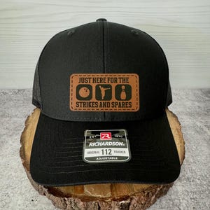 May include: Black trucker hat with a brown leather patch that says "Just here for the strikes and spares" with bowling pins and a bowling ball graphic. The hat is adjustable and has a Richardson logo.