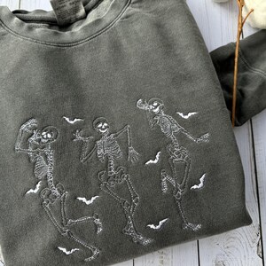 Pode incluir: Uma camisa de moletom verde escura com um design bordado branco de tr&ecirc;s esqueletos dan&ccedil;ando com morcegos voando ao redor deles.