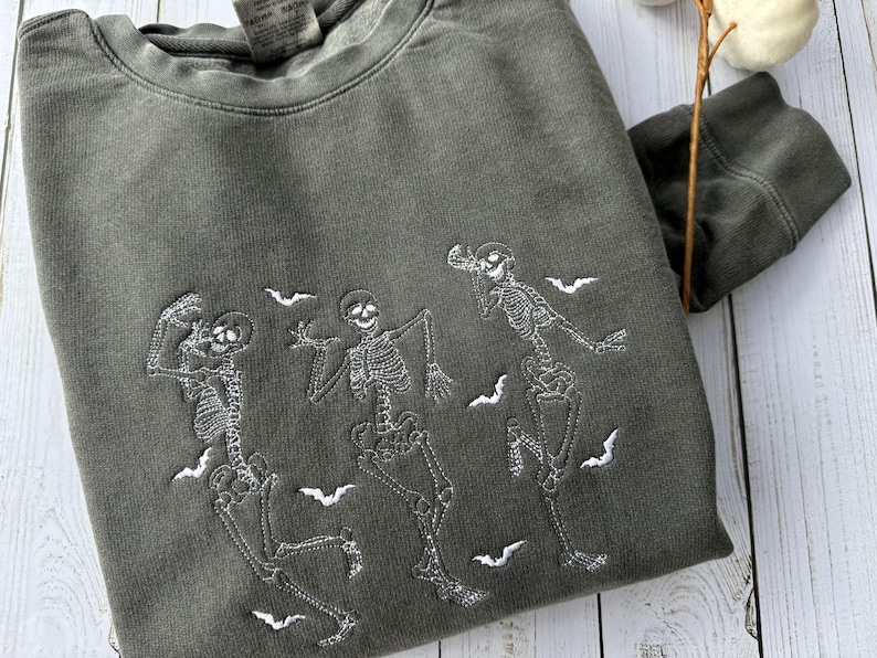 Pode incluir: Uma camisa de moletom verde escura com um design bordado branco de tr&ecirc;s esqueletos dan&ccedil;ando com morcegos voando ao redor deles.