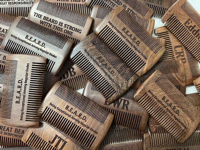 Peut inclure: Gros plan sur un tas de peignes en bois avec le texte "B.E.A.R.D. Being Exceptional Among Regular Dudes" grav&eacute; dessus. Les peignes sont bruns et ont un grain de bois naturel.