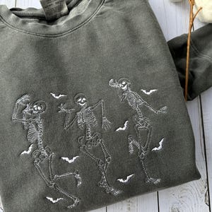 Pode incluir: Uma camisa de moletom verde escura com um design bordado branco de tr&ecirc;s esqueletos dan&ccedil;ando com morcegos voando ao redor deles.