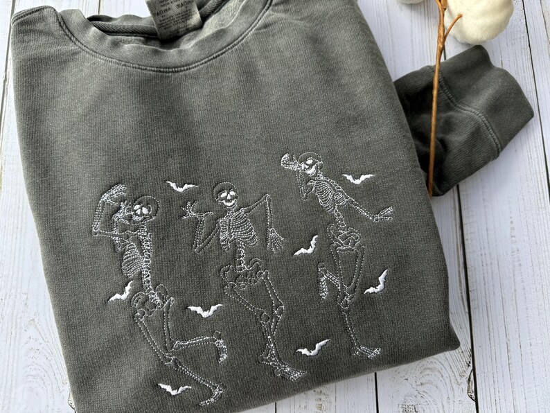 Pode incluir: Uma camisa de moletom verde escura com um design bordado branco de tr&ecirc;s esqueletos dan&ccedil;ando com morcegos voando ao redor deles.