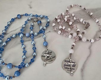 Op de afbeelding: Twee rozenkransen met kralenstrengen. Eén is blauw met kristallen en parelachtige kralen, de andere is roze en wit. Elk heeft een zilveren hanger met een kruis en gegraveerde tekst. De blauwe rozenkrans zegt "Love your Carmelo James 7.5.25". De roze rozenkrans zegt "With love Goddaughter Celina Elise 7.5.25".