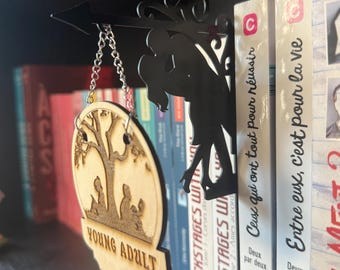 May include: A black metal bookshelf arrow with a silhouette of a couple kissing. A wooden Young Adult sign hangs from the arrow. The sign features a tree and figures. Books are in the background.