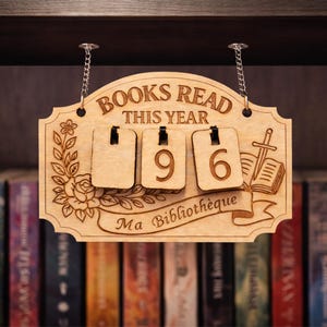 May include: Wooden sign with the words "BOOKS READ THIS YEAR" and the number "96" displayed. The sign has a floral design and the words "Ma Bibliothèque" are written on a banner. The sign is hanging from a chain.