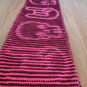 May include: A knitted scarf in pink and burgundy, with the word "GOTH" knitted into the design. The scarf is displayed on a wooden surface, highlighting its textured pattern. The scarf is approximately 150 cm long.