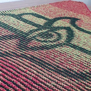 May include: A knitted blanket with a large, dark green trombone design on a yellow and red background. The blanket has a ribbed texture and a red border. This musical-themed blanket is suitable for home decor.