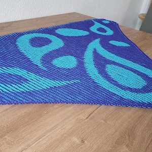 May include: A blue and turquoise knitted shawl with a paisley design. The shawl is triangular, with a dark blue base and a lighter turquoise pattern. The design includes swirls and circular shapes.