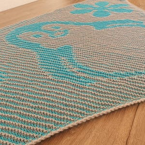 May include: A knitted shawl in a triangular shape, featuring a gray and teal color scheme. The design includes a teal floral pattern and a border. The shawl is placed on a wooden surface.