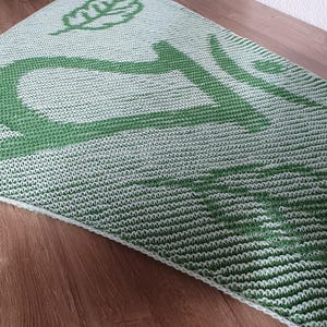 May include: A knitted shawl in shades of green and white, featuring a large green design and a leaf motif. The shawl is triangular and rests on a wooden surface, showcasing its textured pattern and color contrast.