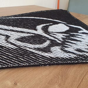 May include: Black and white knitted shawl featuring a skull design. The shawl has a triangular shape with a striped pattern on the edges and a detailed skull graphic in the center. The skull is white, contrasting against the black background.