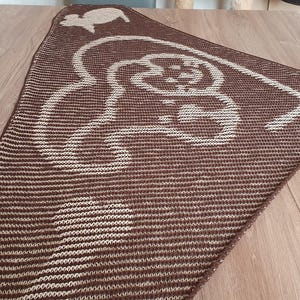 Peut inclure: Un châle tricoté marron et beige avec un motif stylisé de mouton et de fleurs. Le châle triangulaire est posé sur une surface en bois. Le motif beige se détache sur le fond marron.