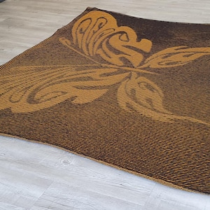 May include: A brown and gold woven rug with a butterfly design. The butterfly is in the center of the rug and is facing to the right.