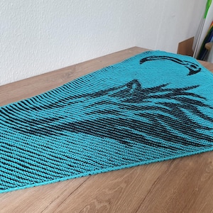 May include: A teal blue knitted shawl with a black design of a crescent moon and stylised leaves. The shawl is made with a textured knit stitch.