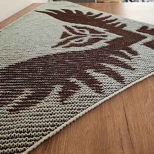 May include: A knitted, triangular shawl featuring a brown bird design against a light gray background. The shawl is displayed on a wooden surface, showcasing the intricate pattern and texture of the knitwear. The bird's wings are spread.