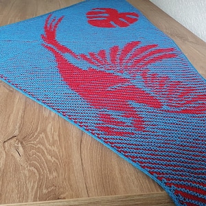 May include: A blue and red knitted shawl with a unique design. The shawl features a large red bird-like figure and a leaf shape on a blue background. The bottom of the shawl has red and blue stripes.