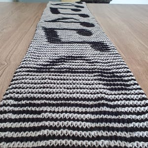 May include: A knitted table runner with a musical theme. The runner is primarily gray with black musical notes and horizontal stripes. The runner is placed on a wooden table.