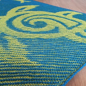 May include: A square, knitted blanket featuring a sea turtle design. The blanket is primarily teal with a lime green turtle motif. The turtle design is centered and takes up most of the blanket's surface. The blanket is placed on a wooden surface.