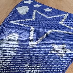 May include: A blue knitted blanket with a large white star in the center and smaller stars and clouds scattered across the top. The blanket is made of a soft, textured material and is likely for a baby or child.