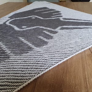 May include: A gray and white knitted shawl with a graphic design of a hand holding a bird. The shawl is folded on a wooden surface, showcasing the intricate knit pattern and the contrasting colors.