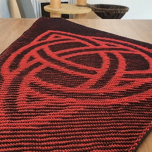 May include: A triangular table runner with a red Celtic knot design on a dark burgundy background. The knitted pattern is displayed, adding a touch of cultural flair to the home. The runner is placed on a wooden table.