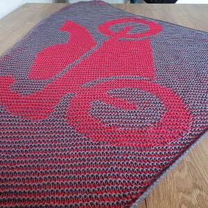 May include: A knitted gray and red blanket with a large red design. The blanket is folded on a wooden surface. The design appears to be a stylized letter or symbol. The blanket has a textured appearance.