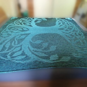 May include: A teal and black throw blanket with a tree of life design. The blanket is folded over a surface, showcasing the intricate pattern. The tree design is centered, with detailed branches and roots.