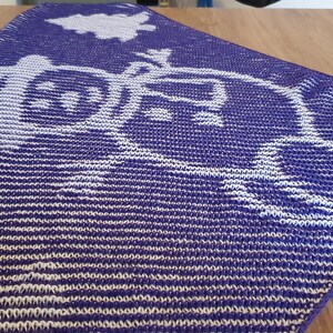 May include: A knitted purple and white blanket with a design of a vehicle and a tree. The blanket has a striped pattern and a detailed image of a vehicle with a tree on top. The blanket is folded on a wooden surface.
