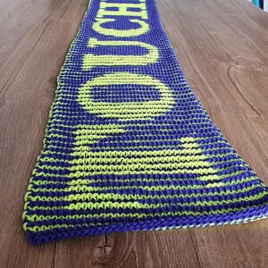 Könnte beinhalten: Ein gestrickter Schal in Blau und Neongrün mit dem Wort "TOUCH" in großen Buchstaben. Der Schal liegt auf einer Holzoberfläche. Der Schal hat eine rechteckige Form.
