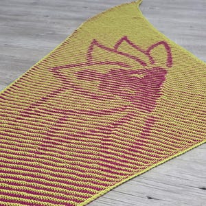 May include: A yellow and magenta knitted shawl with a floral design. The shawl is triangular, with a repeating striped pattern. The floral design is in magenta and is centered on the shawl. The shawl is displayed on a wooden surface.
