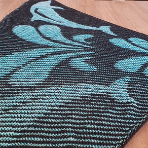 May include: A knitted shawl featuring a dolphin and wave design in teal and black. The shawl is triangular, with a textured, striped pattern. The dolphin is depicted jumping out of the waves.