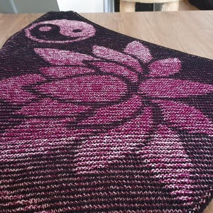 May include: A triangular shawl in shades of black and magenta, featuring a large lotus flower design and a yin-yang symbol. The shawl is made of a knitted fabric and is displayed on a wooden surface.