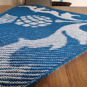 May include: Blue and white knitted table runner with a nautical design. The runner features a repeating pattern of stylized birds, a globe, and waves. The runner is made of a textured knit fabric and is displayed on a wooden table.