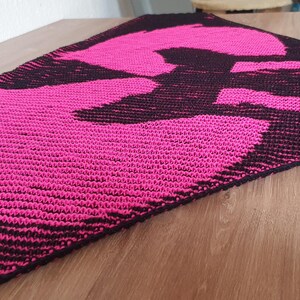 May include: A rectangular, knitted table runner featuring a bold design in black and hot pink. The pattern appears to be an abstract shape. The runner is placed on a light brown wooden surface.