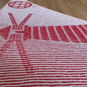 May include: A red and white knitted blanket with a geometric design. The blanket features a repeating pattern of horizontal stripes and a central design with a globe and arrow motif. The blanket is folded on a wooden surface.