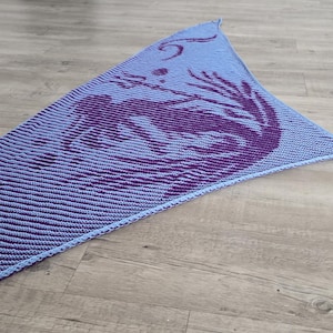 May include: A knitted shawl in shades of blue and purple, featuring a mermaid design. The shawl has a triangular shape with a striped pattern on the lower portion and a detailed mermaid motif near the top. The shawl is lying on a wooden surface.