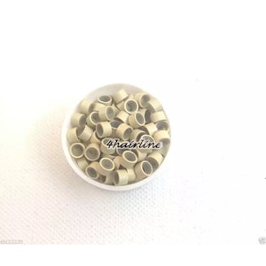 May include: A white bowl filled with numerous small, beige hair extension rings. The rings are cylindrical with a hollow centre. The text "4hairline" is printed on the bowl. The background is white.