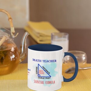 以下が含まれることがあります： ネイビーブルーの内側とハンドルが付いた白いセラミックマグ。マグには「MATH TEACHER」の文字と、「Coffee」、「Patience」、「Chalk Dust」の文字が入った三角形のグラフィックが描かれています。「SURVIVAL FORMULA」というフレーズがグラフィックの下に印刷されています。