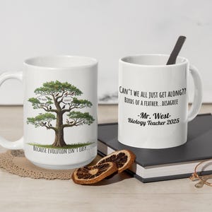 Puede incluir: Dos tazas de cerámica blanca. Una taza presenta una ilustración de un árbol con pájaros y el texto: "BECAUSE EVOLUTION ISN'T EASY...". La otra taza tiene el texto: "CAN'T WE ALL JUST GET ALONG?? BIRDS OF A FEATHER...DISAGREE -Mr. West- Biology Teacher 2025".