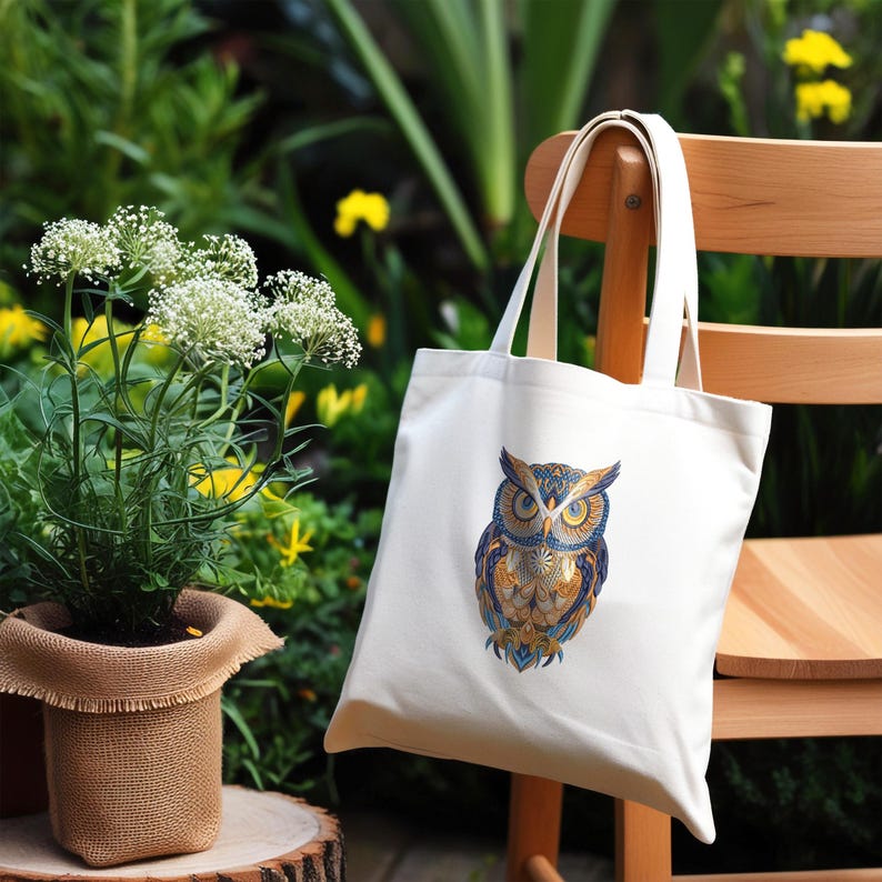 May include: A white tote bag with a detailed owl illustration in shades of blue, gold, and brown. The bag is hanging on a wooden chair, with greenery in the background. The owl design is centered on the bag.