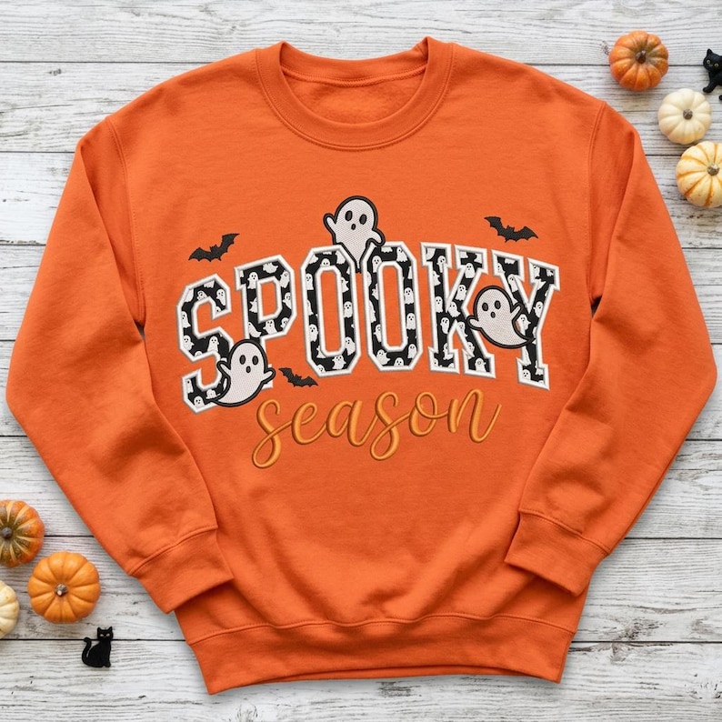 May include: An orange crewneck sweatshirt featuring the text SPOOKY in large block letters with a black and white ghost pattern, and the word season in cursive orange script. Small black bats and white ghost illustrations are included.