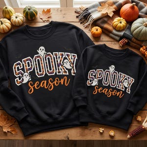 May include: Two black crewneck sweatshirts featuring the text SPOOKY in a plaid orange, black, and white varsity font, with the word season in orange cursive script. Small white ghost illustrations and black bat silhouettes are printed around the text.