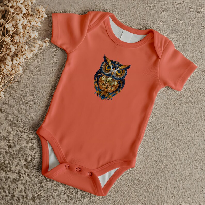May include: Coral-colored baby bodysuit with a detailed owl graphic. The owl features blue, orange, and yellow accents. The bodysuit has short sleeves and a white inner lining. It has snap closures at the bottom.