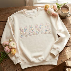May include: A cream-coloured sweatshirt featuring the word "MAMA" in floral print. The sweatshirt is displayed on a wooden surface, with flowers and a mug of coffee. A comfortable and fashionable garment.