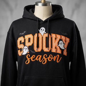May include: A black hooded sweatshirt featuring the embroidered text SPOOKY in orange and white gingham block letters and the word season in orange cursive script. The design includes three white ghost figures and two small black bats.
