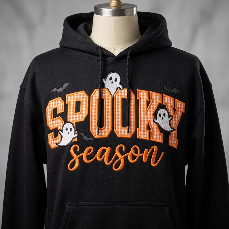 May include: A black hooded sweatshirt featuring the embroidered text SPOOKY in orange and white gingham block letters and the word season in orange cursive script. The design includes three white ghost figures and two small black bats.