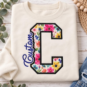 Könnte beinhalten: Ein cremefarbenes Sweatshirt mit Rundhalsausschnitt und einem großen, blockartigen Buchstaben C mit schwarzem Stickrand. Das Innere des Buchstabens ist mit einem bunten Blumenmuster gefüllt. Das Wort Custom ist in blauer Schreibschrift gestickt.