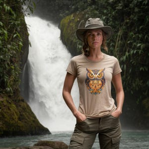 May include: A tan t-shirt featuring a detailed owl design in shades of orange, blue, and yellow. The owl graphic is centered on the front of the shirt. The person is wearing a hat and cargo pants, standing in front of a waterfall.