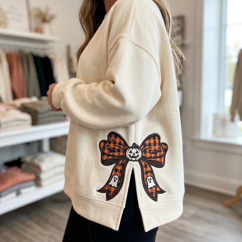 May include: A cream-colored long-sleeve sweatshirt featuring a side slit and a large graphic print of an orange and black plaid bow. The bow design includes a central jack-o'-lantern face, small white ghosts, and tiny star accents on the fabric.